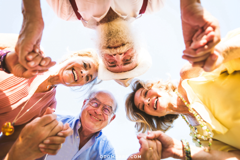 A joyful group of elderly adults stands in a circle, holding hands and smiling widely at the camera. They are outdoors under a clear blue sky, exuding happiness and camaraderie. The website 'ODOMCARE.COM' is displayed at the bottom of the image