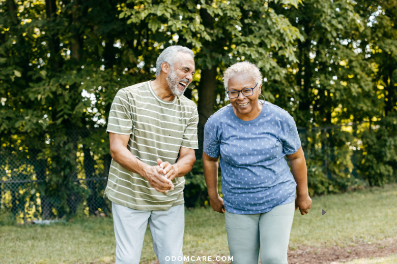 An elderly couple, both with gray hair, laugh and enjoy a moment together outdoors. The man is wearing a green striped shirt and light gray pants, while the woman is dressed in a blue polka dot shirt and light pants. They are in a park-like setting with greenery in the background. The website 'ODOMCARE.COM' is displayed at the bottom of the image.