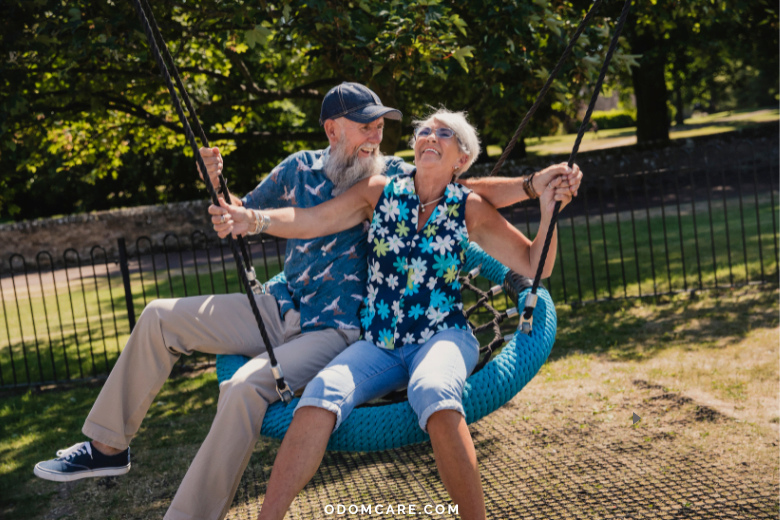 A cheerful elderly couple swings together on a large, round swing in a sunny park. Both are laughing and enjoying the moment, with the man wearing a cap and the woman in sunglasses. The website 'ODOMCARE.COM' is displayed at the bottom of the image.