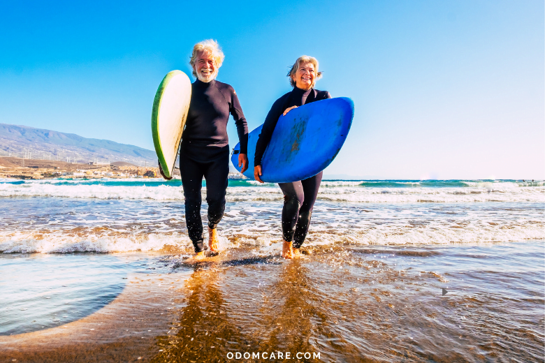 An elderly man and woman in wetsuits walk along the beach, each carrying a surfboard. They are smiling and enjoying a sunny day by the sea. The website 'ODOMCARE.COM' is displayed at the bottom of the image.