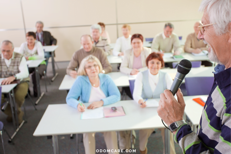An aging adult man with gray hair and glasses holds a microphone while addressing a group of elderly adults seated at desks in a classroom setting. The attendees, both men and women, are attentively listening and taking notes. The atmosphere is lively and engaging, emphasizing the focus on lifelong learning and education. The website 'ODOMCARE.COM' is displayed at the bottom of the image.