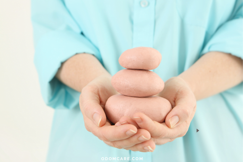 A close-up of a person in a light blue shirt holding a small stack of three smooth, pink stones, symbolizing balance and tranquility. The website 'ODOMCARE.COM' is displayed at the bottom.