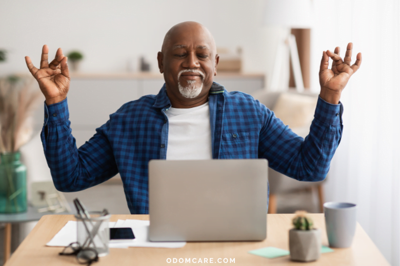 An elderly man with a bald head and a gray beard is sitting at a desk in a modern home setting, meditating with his eyes closed and hands in a yoga mudra pose. He is wearing a blue plaid shirt over a white t-shirt. In front of him is a laptop, along with glasses, a smartphone, papers, and a coffee cup on the desk. The background includes a bookshelf and soft lighting, creating a calm and serene atmosphere. The website 'ODOMCARE.COM' is displayed at the bottom of the image.