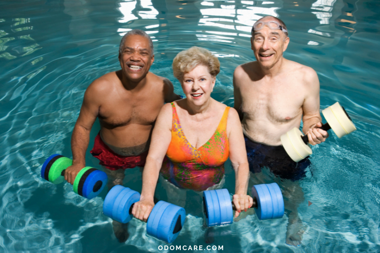 Three cheerful elderly adults are enjoying a water exercise class in a swimming pool. The group includes a man with short black hair and a red swimsuit holding green and blue foam dumbbells, a woman with short blonde hair wearing a colorful swimsuit holding blue foam dumbbells, and a man with a bald head, wearing swim goggles and black swim trunks, holding yellow foam dumbbells. The background is the calm blue water of the pool. At the bottom, the website 'ODOMCARE.COM' is displayed.