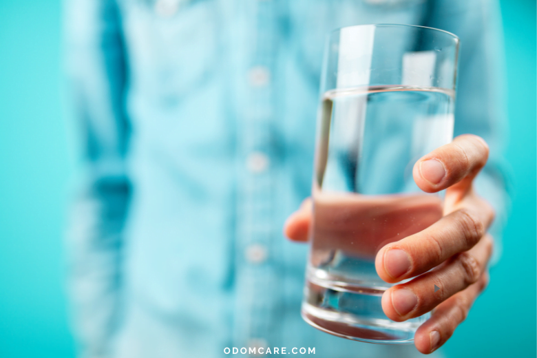 A close-up of a person's hand holding a clear glass of water against a bright turquoise background. The person is wearing a light blue shirt, but their face is not visible. The website 'ODOMCARE.COM' is displayed at the bottom