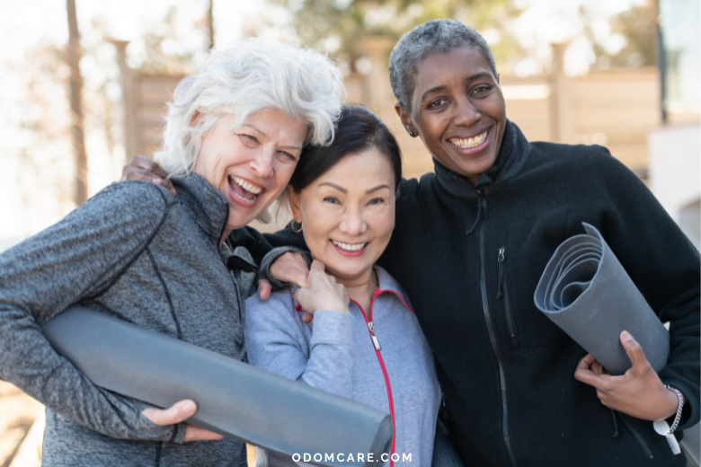 Three aging adult women, each holding a yoga mat, smile and embrace each other outdoors. They are dressed in athletic wear, ready for a fitness session. The background features a sunny, natural setting. The website 'ODOMCARE.COM' is displayed at the bottom.