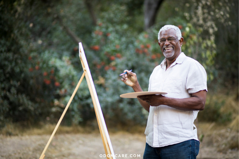 An aging adult man with gray hair and glasses, dressed in a light short-sleeve shirt, is standing outdoors with an easel, painting. He is smiling broadly, enjoying his creative activity in a natural setting. The website 'ODOMCARE.COM' is displayed at the bottom.