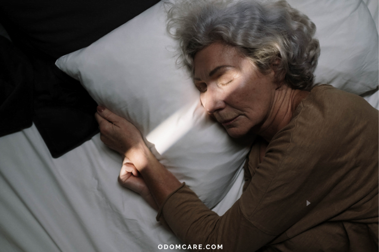 An aging adult woman with gray hair sleeping peacefully on a white pillow in a dimly lit room. A beam of light falls across her face, emphasizing tranquility. The website 'ODOMCARE.COM' is displayed at the bottom.