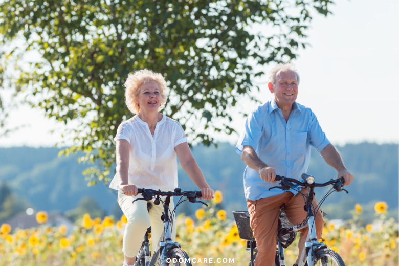 An aging adult couple, a woman and a man, are riding bicycles outdoors on a sunny day. They are both smiling and enjoying the ride, surrounded by a picturesque landscape with sunflowers and trees. The website 'ODOMCARE.COM' is displayed at the bottom